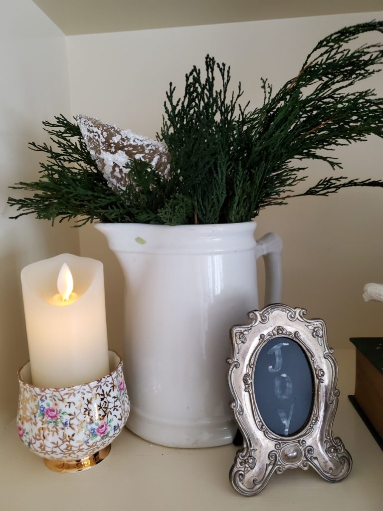 close up of a pitcher with cedar branches a candle and a small frame that says Joy