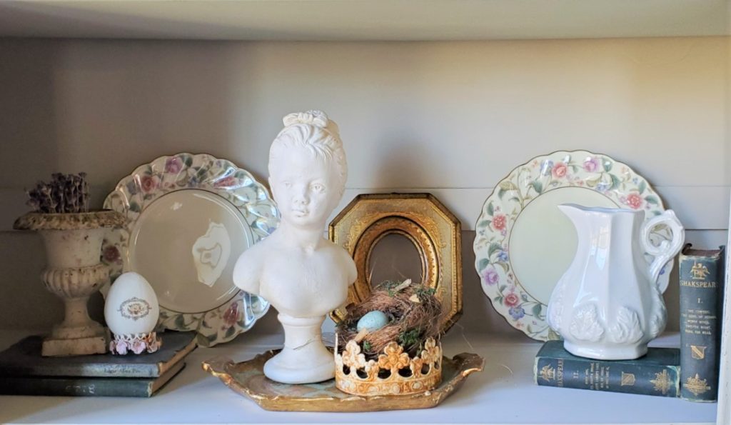 a shelf with a female bust, vintage dishes, crown and bird nest