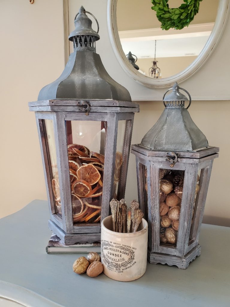 Two lanterns filled with orange slices, nuts and pinecones.  A white dish filled with silverware
