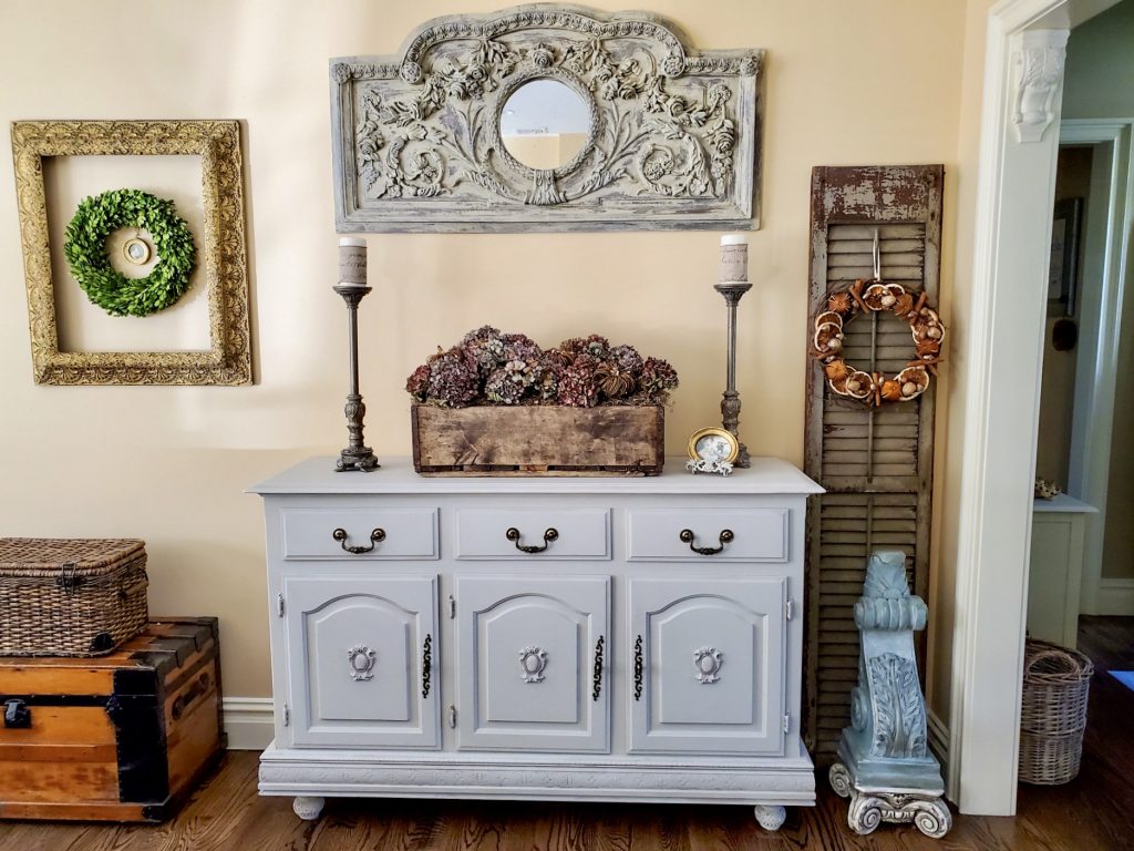 picture of natrual fall decor on the buffet in the great room.
Dry hydrangeas fill a wooden crate with a basket and old steamer trunk next to it.
An old shutter with a corbel on a pedestal under the corbel
a wreath hangs on the shutter
a large mirror hangs over the buffet