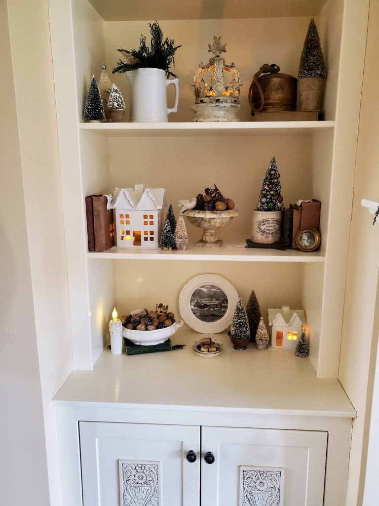 picture of a bookcase with books, houses, candles and bottle brush trees