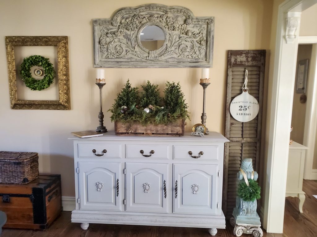 a full view of the buffet with a wood box filled with greens. Candle sticks with candles.
A shutter with a Christmas dign and corbel with a small green wreath