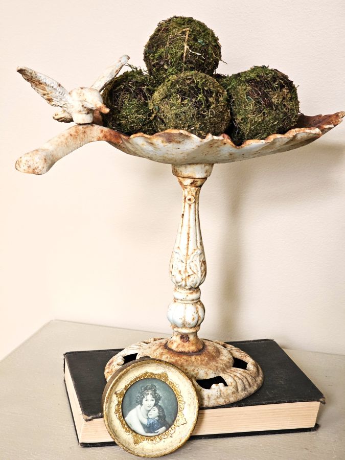 don't-miss-out-on-easy-spring-decorating-ideas-closer-look-at-moss-balls-in-cast-iron-bird-feeder-sitting-on-a-book-with-a-small-picture-featured-image